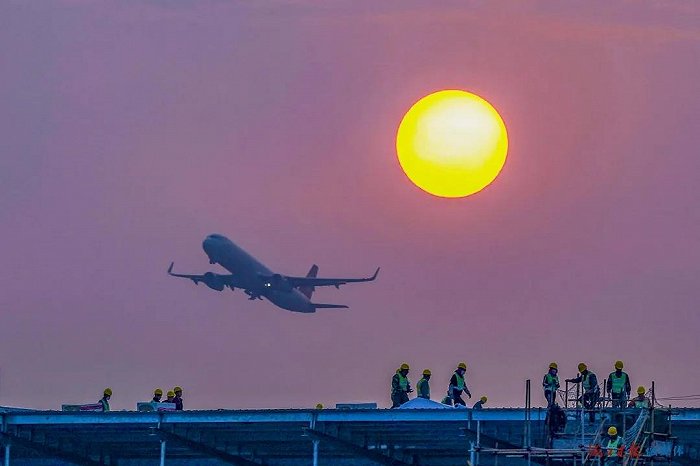 夕阳西下，海口美兰空港一站式飞机维修基地（一期）项目现场一片繁忙，工人正忙着搭建屋面板，一架刚起飞的客机从上空飞过。图片来源：王程龙/摄
