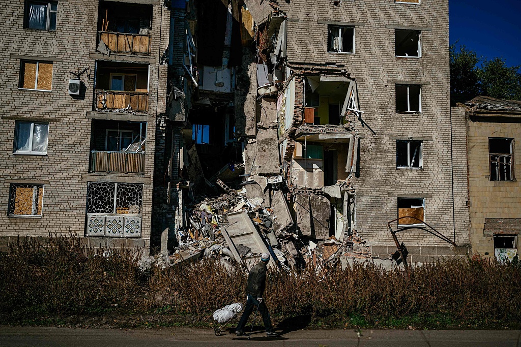 当地时间2022年10月15日，顿涅茨克地区巴赫穆特，一名当地居民推着一辆手推车经过一座受损的建筑。人民视觉 图