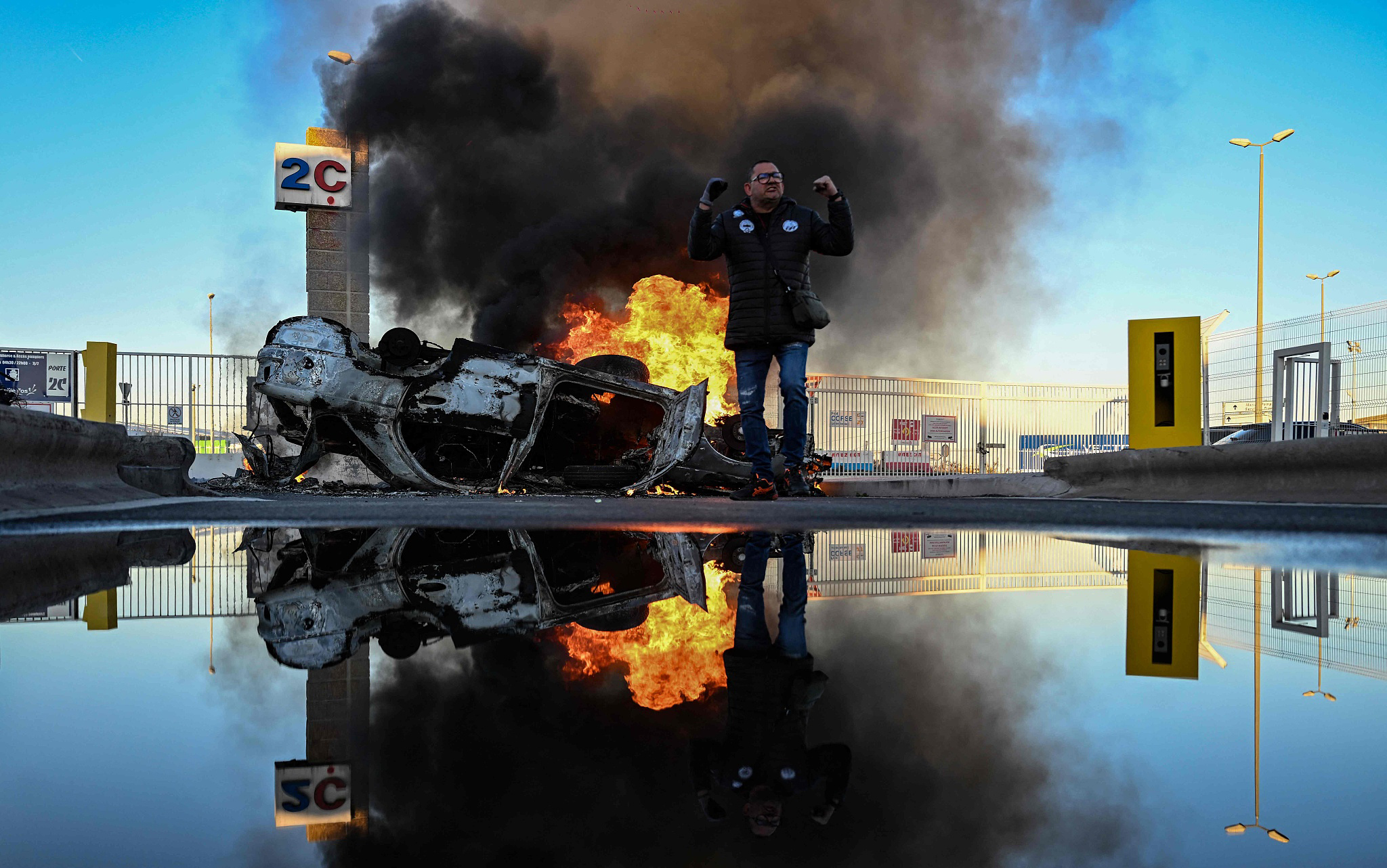 当地时间2023年3月22日，法国南部马赛港，法国劳工联合会成员站在一辆被掀翻的燃烧的汽车前举起手臂。