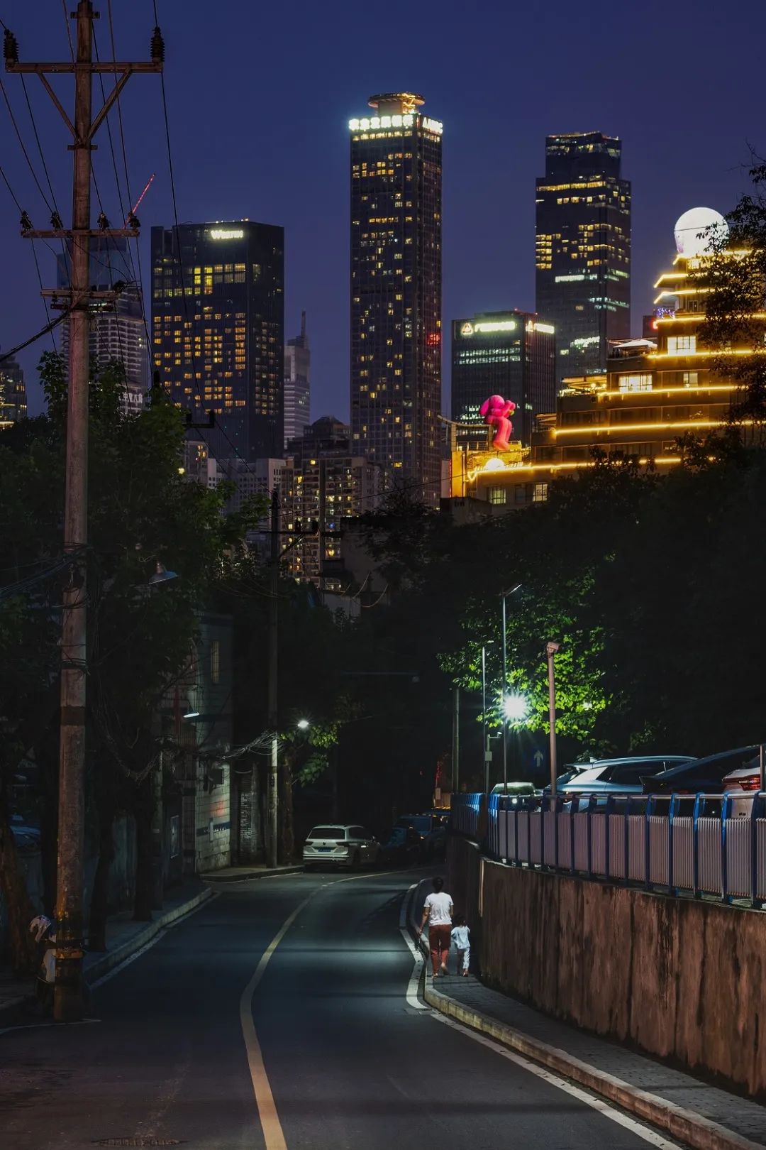 夜晚,远处灯火辉煌的高楼大厦,近处的路灯,行走在人行道上的母女,这就