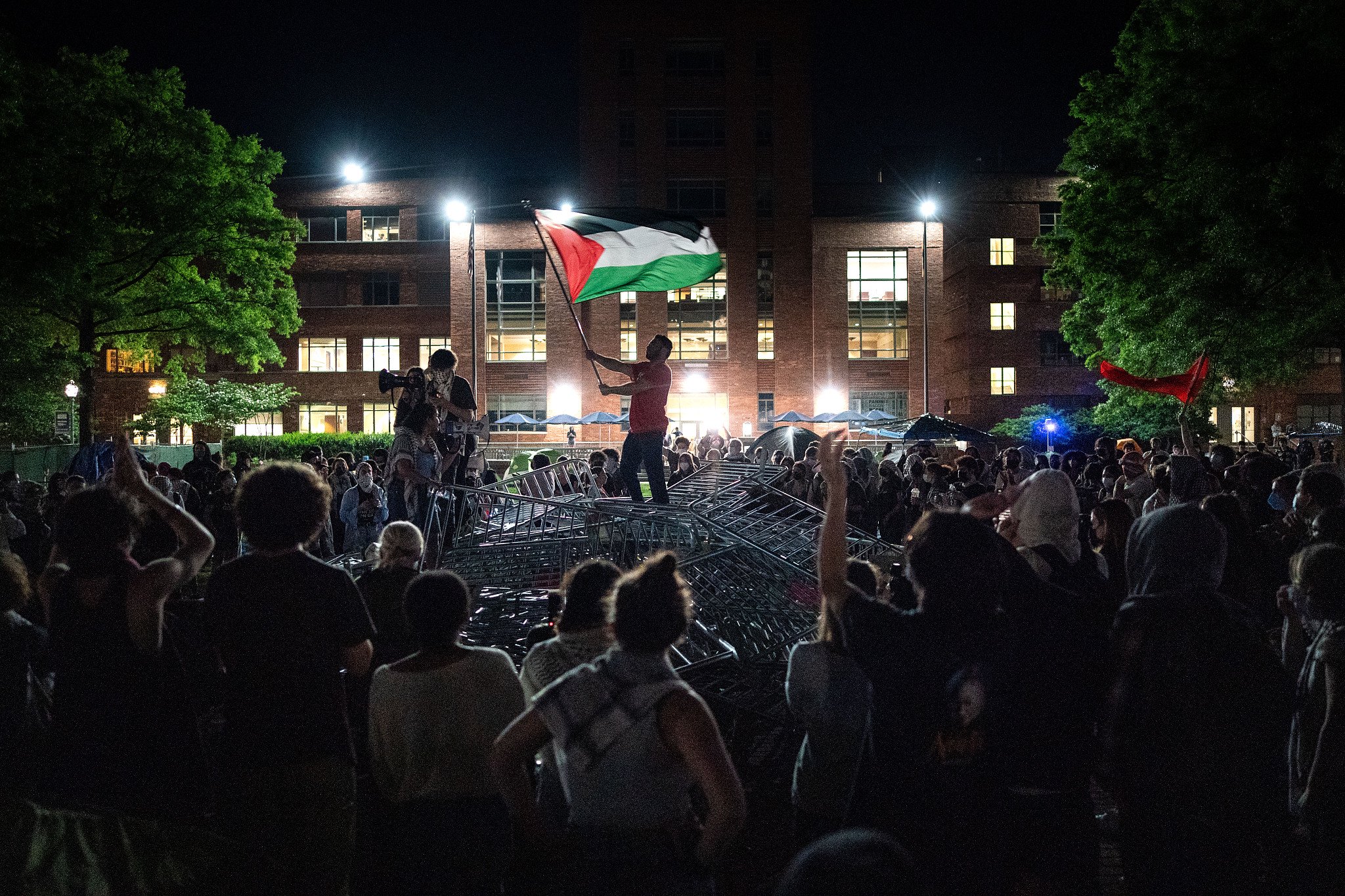 当地时间2024年4月29日，美国华盛顿特区，乔治·华盛顿大学里，一名男子高举一面巴勒斯坦国旗，与学生们一起高呼口号。