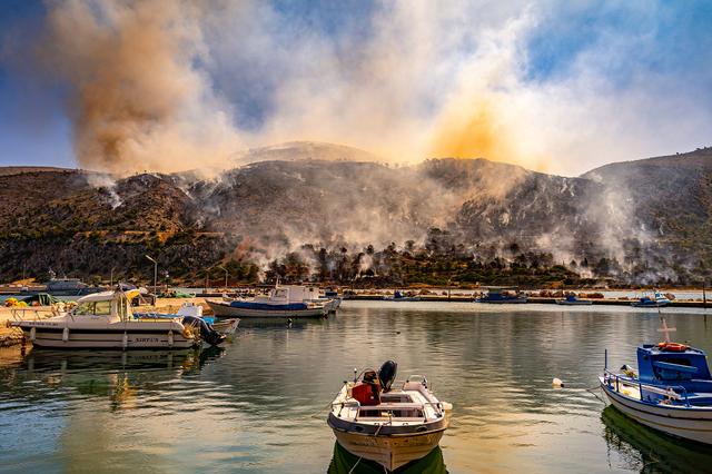 当地时间2025年6月24日，希腊希俄斯岛，渔船停泊在港口，背景是森林地区燃起的野火。视觉中国 图