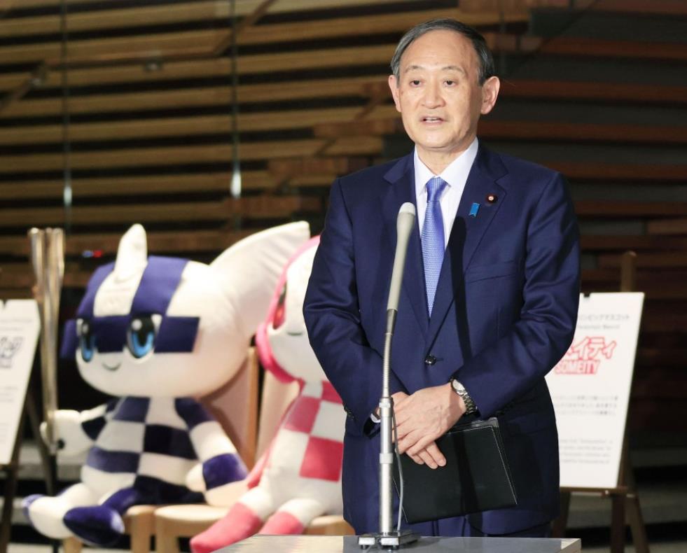 △2020年12月31日，日本首相菅义伟在东京办公室与新闻记者谈话，身后两个人偶均为2020东京奥运会吉祥物。（图片来源：共同社）