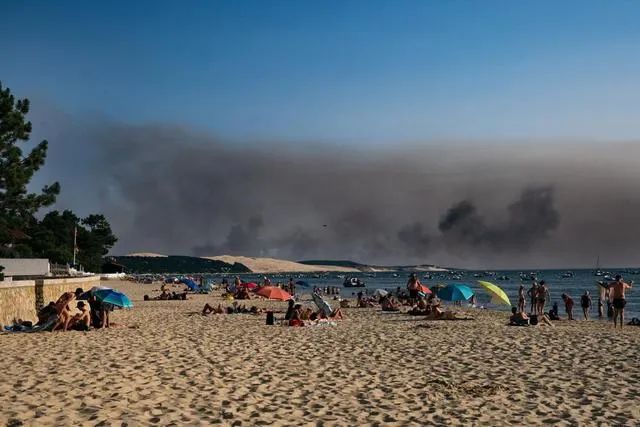当地时间2022年7月16日，法国西南部兰蒂拉斯，游客在海滩避暑，远处森林大火产生的黑色浓雾在步步逼近
