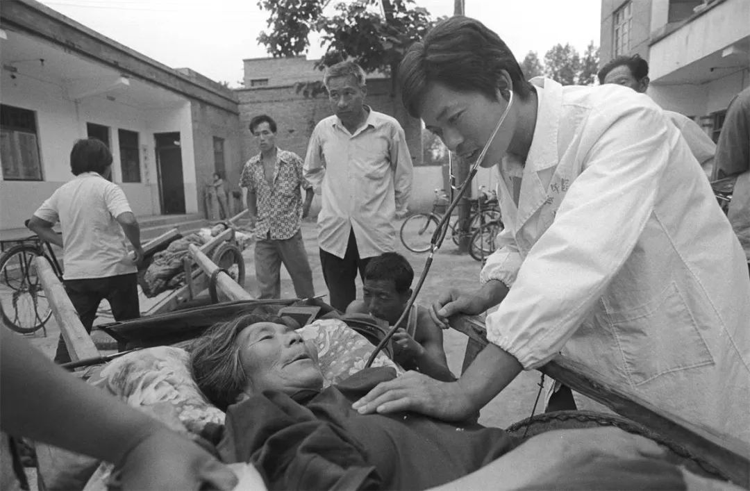 1996 年，河南宜阳，村民拉着架子车送家人到乡卫生院看病，医生查看病人病情 （吕光伟 摄/ 视觉中国 供图）